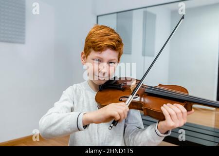 Fröhliches Ingwer-Kind spielt Geige und schaut auf die Kamera, während es beim Musikunterricht Melodie lernt Stockfoto