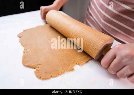 Draufsicht auf die Hände einer Frau, die den Teig mit einem hölzernen Nudelholz für Kekse aus dem Mürbeteig ausrollen. Stockfoto