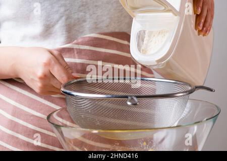 Nahaufnahme Frau siebt Weizenmehl durch ein Sieb in eine Rührschüssel zur Teigvorbereitung für Kuchen und Backwaren auf schwarzem Hintergrund. Den Teig für hausgemachtes Gebäck kneten. Stockfoto