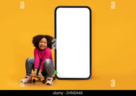 Fröhliches kleines schwarzes Mädchen, das auf dem Skateboard in der Nähe eines riesigen Smartphones mit leerem weißen Bildschirm sitzt, Mockup Stockfoto