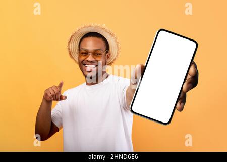 Aufgeregt schwarzer Mann im Sommer Hut zeigt Handy mit leerem Bildschirm, empfehlen mobile App für Reisen Stockfoto