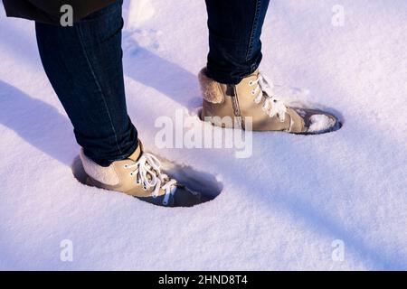 Nahaufnahme von Frauenbeinen in hellem Wildleder und blauen Jeans, die an einem sonnigen Wintertag im Schnee stehen. Winterspaziergang an der frischen Luft Stockfoto