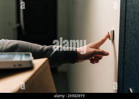Nahaufnahme der Hände eines nicht erkennbaren Zustellers, der die Türklingel der Kundenwohnung mit einem Karton und einem POS-Terminal für kontaktlose Zahlungen anklingelt. Stockfoto