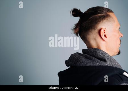 Ein Nahaufnahme-Foto eines jungen Mannes in einer Weste an der Seite. Ein Mann mit langen Haaren, die in einem Knoten oder einem Männerbrötchen zusammengetragen sind. Auf blauem Hintergrund sind Männerfrisuren, Stockfoto