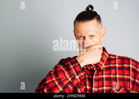 Ein Nahaufnahme-Foto eines ernsthaft nachdenklichen jungen Mannes in einem roten karierten Hemd. Ein Mann mit langen Haaren, die in einem Knoten zusammengeknotet sind, und mit der Faust auf die Wange stanzt, Stockfoto