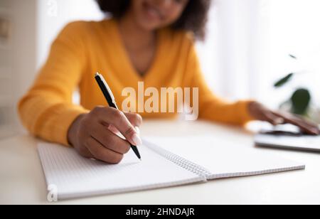 Nicht erkennbare junge schwarze Frau, die während des Online-Unterrichts auf dem Laptop von zu Hause aus Nahaufnahme Informationen im Notebook aufschreibt Stockfoto