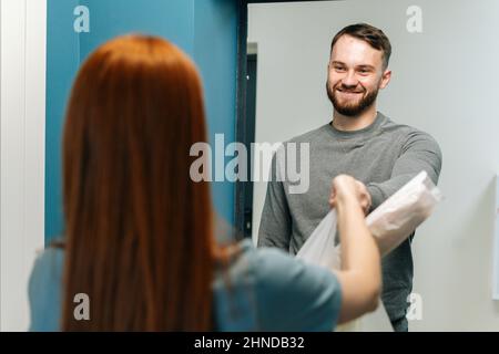 Fröhlicher bärtiger Kurier männlich liefert Lebensmittelbeutel mit zu unkenntlich weiblichen Hausfrau Kunde auf Tür zu Hause. Stockfoto