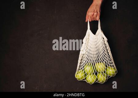 Eine Frauenhand hält einen Saitenbeutel mit grünen Äpfeln auf einem dunklen strukturierten Hintergrund, das Konzept der abfallfreien Produktion, die Ablehnung von Plastiktüten Stockfoto