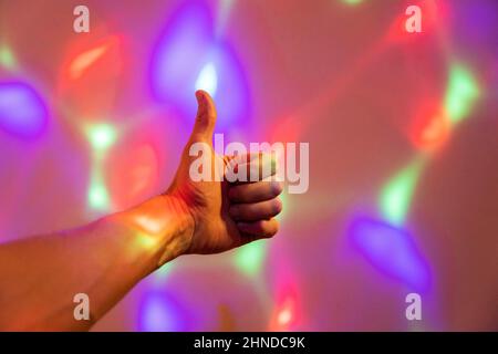 Die Hand des Mannes gibt einen Daumen nach oben, vor dem Hintergrund der laufenden farbigen Highlights, Disco und Lichtshow, Weihnachtslichter, farbige Running Christ Stockfoto