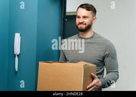 Porträt eines freundlichen bärtigen Kurierers, der das Pappkartonpaket in den Händen hält, die am Eingang der Kundenwohnung stehen. Stockfoto