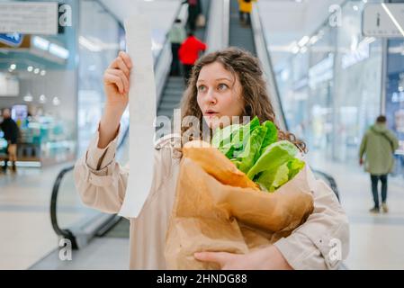 Steigende Lebensmittelpreise. Überraschte Frau, die im Einkaufszentrum in einen Papierscheck schaute und eine Papiertüte mit frischen Kräutern und einem Baguette in der Hand hielt. Stockfoto