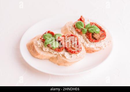 Bruschettas oder Toasts mit Ricotta-Käse und in Scheiben geschnittenen sonnengetrockneten Tomaten, gekrönt mit Basilikum auf weißem Teller, weißer Tischhintergrund. Köstlicher Klassiker Stockfoto