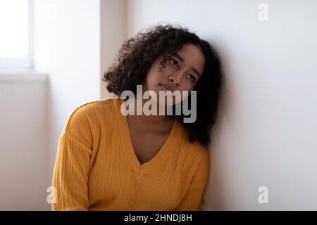 Depressive junge schwarze Frau, die allein in der Nähe der Wand sitzt und zu Hause ein psychisches oder psychologisches Problem hat Stockfoto