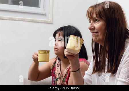 Glückliche lateinamerikanische argentinische Mutter und Tochter, die zu Hause sitzen und Kaffee trinken und Fernsehen, zusammen genießen, junge Frau mit Down-Syndrom, die ein c Stockfoto
