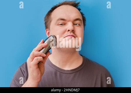 Porträt eines zerzausten erwachsenen Mannes, der mit einem elektrischen Rasiermesser Stoppeln rasiert, blauer Studiohintergrund Stockfoto