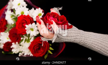 Frauenhand mit roter Rosenknospe. Weibliche Hand in stilvollem Strickhandschuh mit Blumenkopf auf schwarzem Hintergrund mit Blumenstrauß Stockfoto