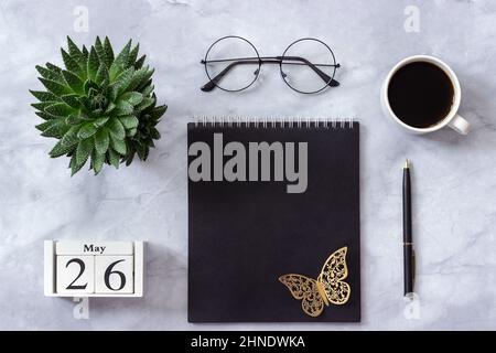 Schreibtisch im Büro oder zu Hause. Holzwürfel Kalender Mai 26. Schwarzer Notizblock, eine Tasse Kaffee, Sukkkulent, Gläser auf Marmorhintergrund Konzept stilvolle Workpla Stockfoto