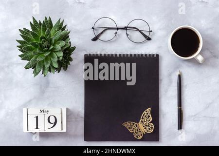 Schreibtisch im Büro oder zu Hause. Holzwürfel Kalender Mai 19. Schwarzer Notizblock, eine Tasse Kaffee, Sukkkulent, Gläser auf Marmorhintergrund Konzept stilvolle Workpla Stockfoto