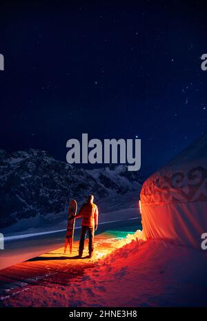 Mann mit geteiltem Snowboard in der Nähe des nomadischen Wohnkomplexes Yurt im Skigebiet Shymbulak in Almaty, Kasachstan. Winternacht Astrofotografie mit Sternen A Stockfoto