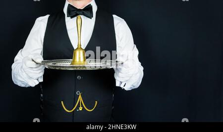 Butler oder Kellner in schwarzer Weste und weißen Handschuhen mit Goldglocke auf silbernem Tablett. Ring für Servicekonzept. Professionelle Gastfreundlichkeit. Stockfoto