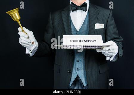 Butler in formellem Anzug und weißen Handschuhen mit goldener Glocke und silbernem Schild. Klingeln für Service. Konzept der professionellen Gastfreundschaft. Stockfoto