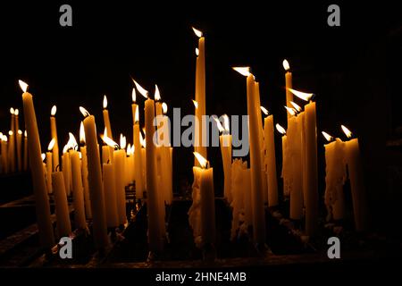 Nahaufnahme der Gruppe weiße gelbe Wachskerzen, die im Wind brennen, dunkelschwarzer Hintergrund in der Kirche Stockfoto