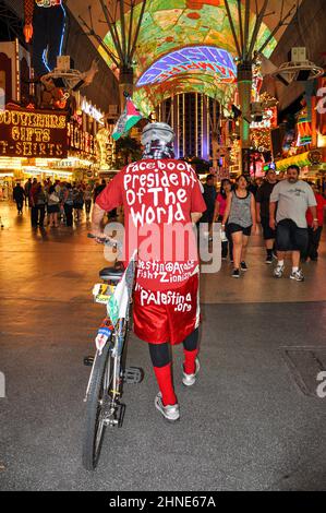 Ein Mann in exzentrischer Kleidung schiebt auf der Fremont Street in Las Vegas ein Fahrrad. Er trägt rote Shorts, Socken, Hemd, Botschaften in weißen Buchstaben. Stockfoto