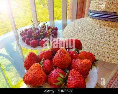 Eine Nahaufnahme eines Tellers mit Erdbeeren, ein Strohhut auf einer Terrasse im Freien an einem sonnigen Sommertag, das Konzept eines luxuriösen Sommerurlaubs wi Stockfoto