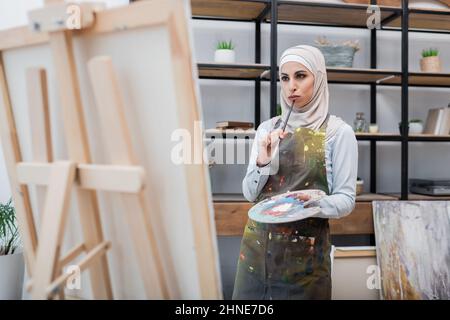 Nachdenkliche arabische Frau mit Palette und Pinsel, die zu Hause fast an eine Staffelei denkt Stockfoto