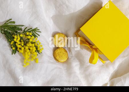 Zweige gelber Mimosenblüten auf weißen Leinenbettlaken, französischen gelben Makronen und gelber Geschenkbox. Gruß zum Internationalen Frauentag am März Stockfoto