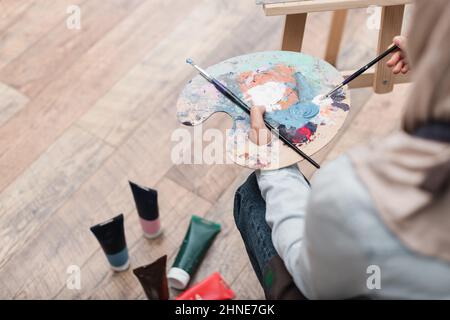Blickfeld einer verschwommenen Frau mit Palette und Pinsel in der Nähe von Farbschläuchen auf dem Boden Stockfoto