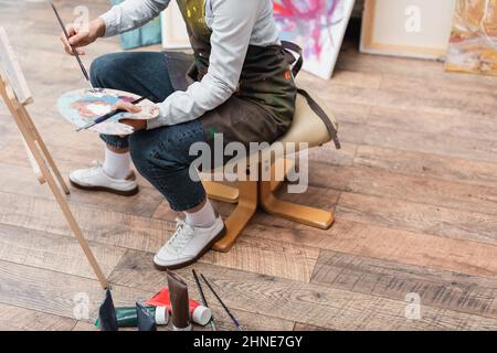 Teilansicht einer Frau mit Palette und Pinsel, die auf einem Stuhl in der Nähe einer Staffelei sitzt Stockfoto
