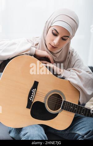 Junge arabische Frau in Hijab, die sich zu Hause auf akustische Gitarre stützt Stockfoto