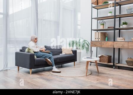 eine arabische Frau mit einer Schüssel Popcorn, die sich im modernen Wohnzimmer einen Film auf einem Laptop ansieht Stockfoto