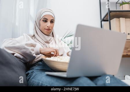 Positive Mulsim-Frau mit einer Schüssel Popcorn, die sich zu Hause auf einem verschwommenen Laptop einen Film ansieht Stockfoto