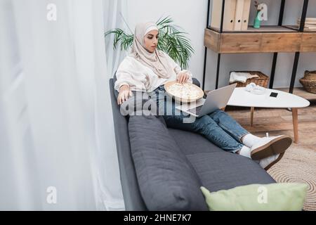 In voller Länge sehen Sie eine arabische Frau in einem Hijab, die mit Popcorn auf dem Sofa sitzt und sich am Computer einen Film ansieht Stockfoto