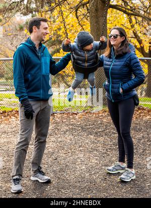 Junge Eltern schieben den zweijährigen Jungen auf eine Schaukel auf dem Spielplatz der Stadt; Philadelphia; Pennsylvania; USA Stockfoto