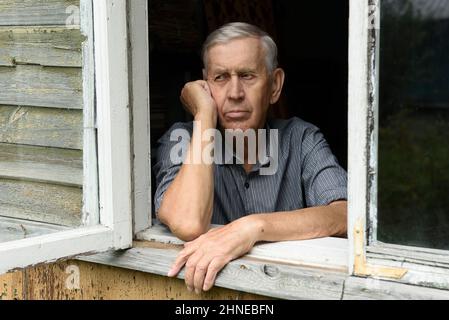Porträt eines traurigen älteren Mannes von 80 Jahren, der im Fenster eines alten Holzhauses sitzt. Stockfoto