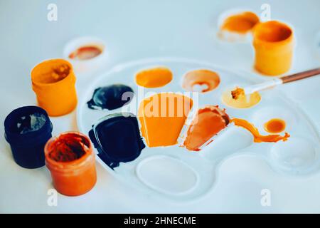 Auf dem weißen Tisch befindet sich die weiße Farbpalette, daneben befinden sich die Gläser der Gouache verschiedener Farben und der Pinsel der Farbe. Kreativ Stockfoto