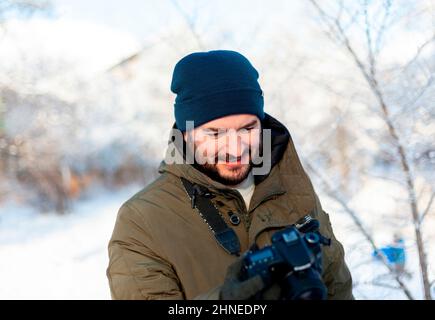 Ein bärtiger Mann in einem Winterhut und Kleidung hält eine Kamera in Fäustlingen und blickt auf seinen Bildschirm. Fotograf bei Fotoshootings im Winter im Freien Stockfoto