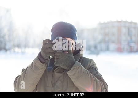 Ein Mann mit Wintermütze und Fäustlingen schießt mit einer digitalen Spiegelreflexkamera, während er im Freien steht. Fotoshooting während der Weihnachtsfeiertage Stockfoto