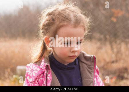 Ein kleines schönes, niedliches Mädchen in einer Jacke mit blondem Haar in Zöpfen steht auf einem herbstlichen Hintergrund und blickt traurig nach unten Stockfoto