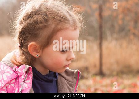 Kleine schöne niedliche traurige Mädchen in einer Jacke mit blonden Haaren in Zöpfen steht auf einem Herbst Hintergrund Stockfoto