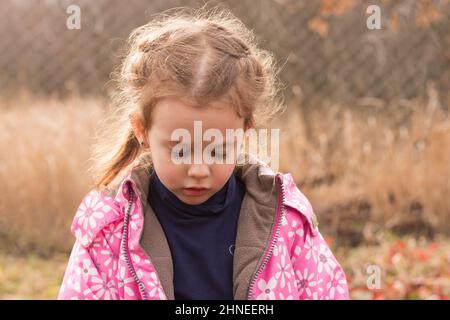 Kleine schöne niedliche traurige Mädchen in einer Jacke mit blonden Haaren in Zöpfen steht auf einem Herbst Hintergrund Stockfoto