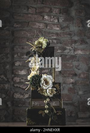 Dreistufige quadratische Hochzeitstorte mit Blumen und dunkelgrünen Akzenten auf Steinwand Hintergrund Stockfoto