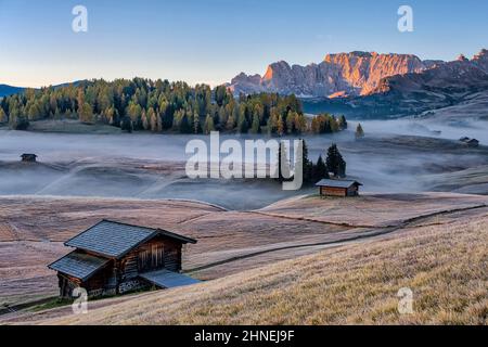 Nebel über den Weiden der Seiser Alm mit Holzhütten, die Roterdspitze in der Ferne, im Herbst bei Sonnenaufgang. Stockfoto