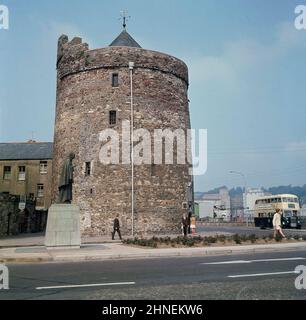 1960s, Blick aus dieser Zeit auf den historischen Reginald's Tower in Waterford, Irland. Ein antikes Gebäude, teilweise aus der Zeit der Wikinger, wurde es im 13.. Jahrhundert von den Anglo-Normannen grundlegend umgebaut und später wurden im 15.. Jahrhundert zwei obere Stockwerke hinzugefügt. Es befindet sich im Kayside-Bereich, bekannt als das Wikingerdreieck. Ein blau-cremefarbener Doppeldeckerbus des Tages ist mit einer Werbung für das Powers Gold Label zu sehen. Stockfoto