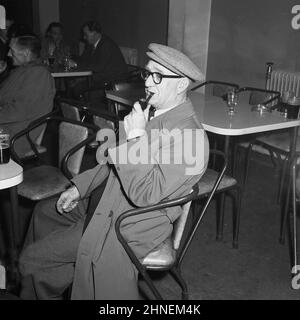 1950s, historisch, ein Arbeiter, der Jacke und Hose, Regenmantel, Mütze und Pfeife raucht, bei einem Drink und einem Pint Bier im sozialen Club des Unternehmens nach der Arbeit, Port Talbot, Wales, Großbritannien, sitzt. Stockfoto