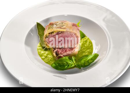 Gebackenes Schweinefilet, mit Speck auf Erbsencreme pochiert, mit Basilikumblättern auf weißem Teller auf weißem Teller Stockfoto