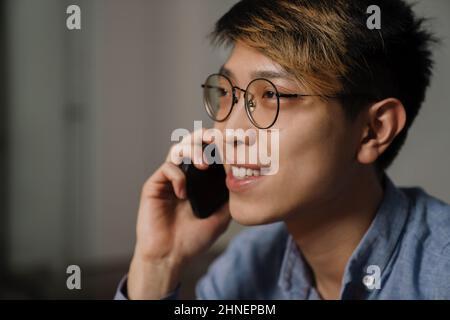 Glücklicher asiatischer Kerl in Brillen, der während der Arbeit im Büro auf dem Mobiltelefon spricht Stockfoto